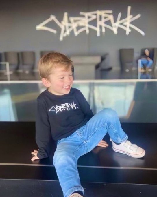 The image features a young boy sitting on a black surface, wearing a black shirt with white text that reads "Cybertrick". He has blue jeans on and is smiling at the camera. The background of the photo shows a room filled with chairs and a sign that says "Cybertrick", indicating that this might be an event or gathering related to the cryptocurrency world.

The post includes a reference to the cryptocurrency "Dogecoin" and a reference to the term "Cybertrick". These elements suggest that the image could be related to a social media post, possibly promoting a cryptocurrency event or simply discussing the concept of "Cybertrick" in general.