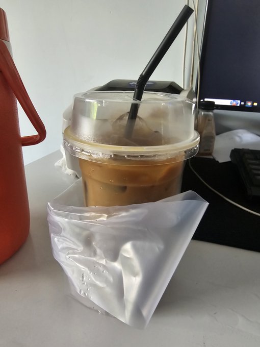 An iced coffee in a clear plastic cup with a black straw, wrapped partially in a white plastic bag, sits on a desk next to a red water bottle and a computer monitor.