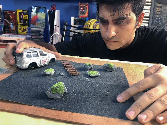 Photograph of a young man leaning over a diorama featuring a white SUV model on a dark surface with miniature rocks, moss patches, and a small wooden ladder, with collectible boxes and a wrestling belt visible on shelves in the background.