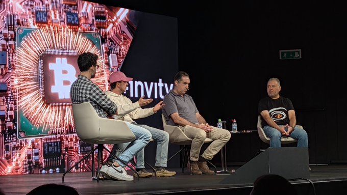A panel discussion on stage at a Bitcoin event with a large Bitcoin logo display and 'infinity' text visible in the background, featuring four seated speakers.