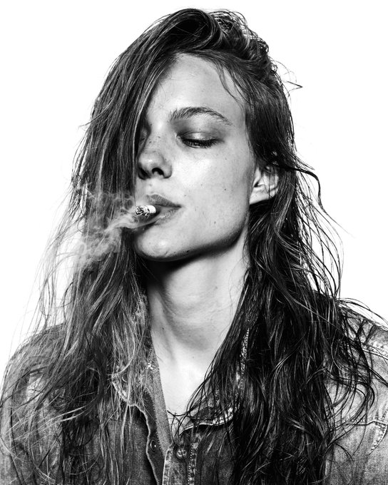A black and white portrait of a young woman with long, wet, tousled hair, eyes closed, smoking a cigarette with smoke curling around her face.