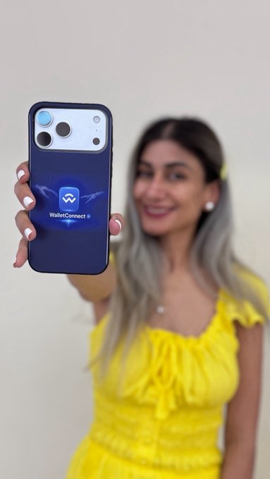 A woman in a yellow dress holds up a smartphone displaying the WalletConnect logo on its screen.