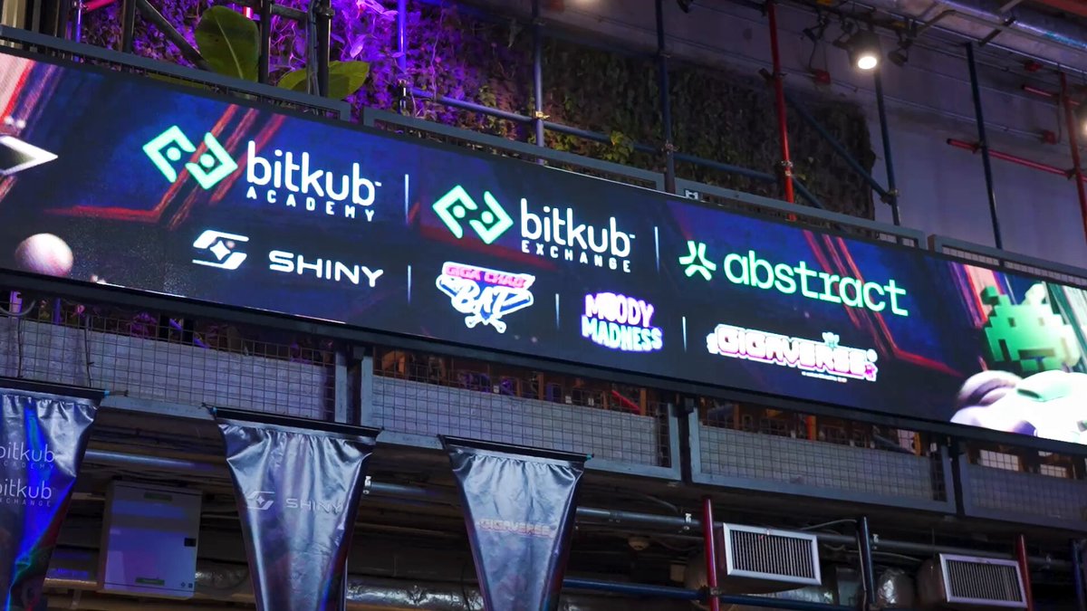 The image shows a large sign with various logos and text that reads "Bitkub Academy" and "abstactract", indicating an event related to Bitkub Academy or a similar organization. The sign also displays the names of some sponsors, such as "Shiny", "Brightness", and "Abstraction". The background of the image is dark, which contrasts with the brightly lit sign and makes it stand out.
