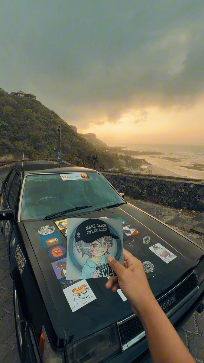 A hand holds an anime-style sticker reading 'MAKE ALIENS GREAT AGAIN' in front of a sticker-covered car hood, with a scenic sunset over a coastal road and mountains in the background.