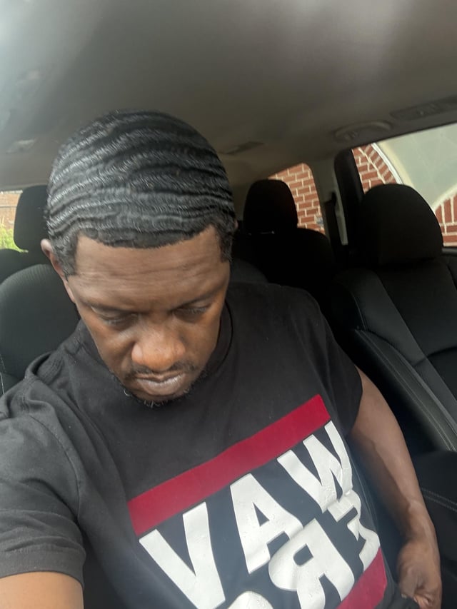 A man sits inside a car looking downward with a somber expression, wearing a black t-shirt with bold red and white graphics reading "VAN" and "85." The contemplative pose and reference to r/Hedera suggest a weary reflection on a slow-moving or stagnant situation, mirroring the frustrations often expressed in that subreddit.