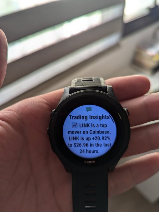 A Garmin smartwatch displaying a Trading Insights notification on a blue screen that reads: 'LINK is a top mover on Coinbase. LINK is up +20.92% to $26.96 in the last 24 hours.'