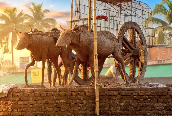 In the image, there are two brown cows standing next to each other near a wall. One cow is facing the camera while the other one is looking away. The background features a street with several buildings. On top of these buildings, there's an advertisement for Nomisma network super bullish @useTria or @perx_trade, which indicates that the image might be related to some form of advertisement or promotion involving cows and the network platform.