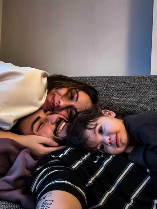 Three young men are huddled together smiling and laughing while taking a selfie on a gray couch.