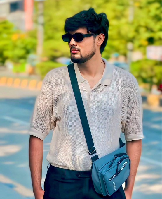 A young man with dark hair wearing black sunglasses, a beige knit polo shirt, and a blue crossbody bag standing outdoors on a sunny street.
