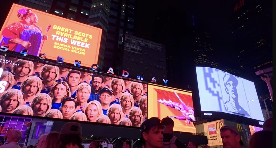 A nighttime photo of illuminated billboards in Times Square, including an advertisement for "Buena Vista Social Club" reading "GREAT SEATS AVAILABLE THIS WEEK" and a large screen displaying a crowd of blonde men with the word "BROADWAY."