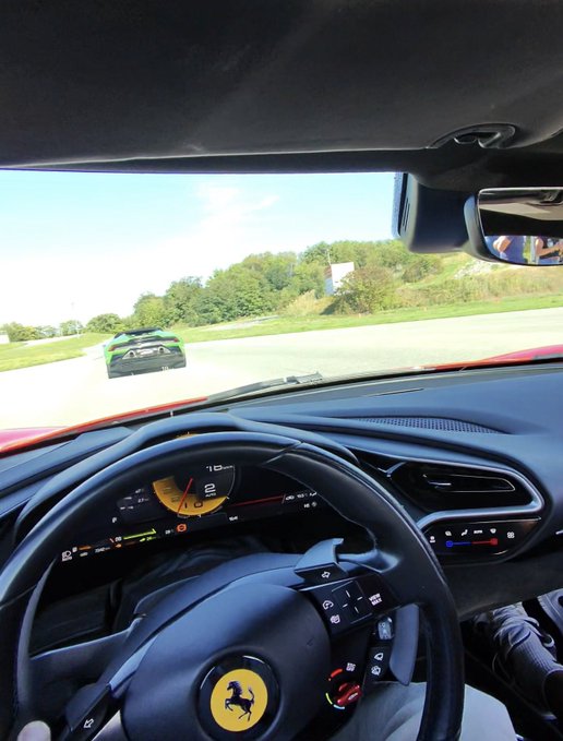 A view from inside a Ferrari, showing the steering wheel with the prancing horse logo, the dashboard, and a green sports car driving ahead on a track or road.