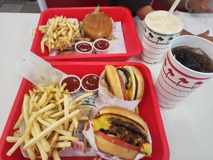 Two red trays on a table hold burgers, fries, and small cups of ketchup, alongside a milkshake and a soda.