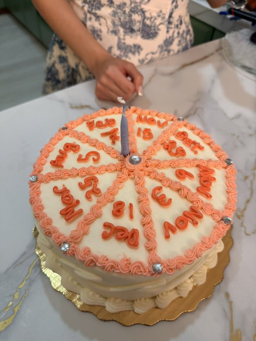 A birthday cake decorated with pink frosting borders and silver accents has a single lit candle in the center, surrounded by written months and numbers indicating an early date.