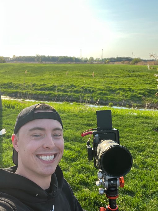 A smiling young man wearing a black cap and hoodie takes a selfie in a grassy field while holding professional camera equipment on a tripod.