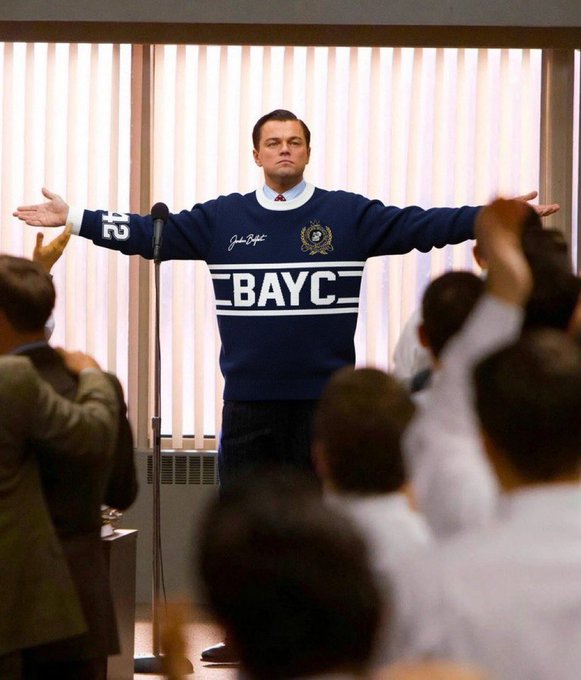 Leonardo DiCaprio in a scene from The Wolf of Wall Street standing with arms outstretched, digitally edited to wear a blue sweater with 'BAYC' and the number 25, with an audience cheering around him.