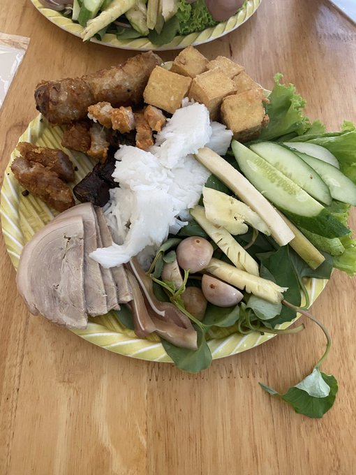 A yellow plate filled with a variety of Vietnamese food, including rice noodles, sliced pork, fried tofu, egg rolls, cucumber, and fresh herbs.