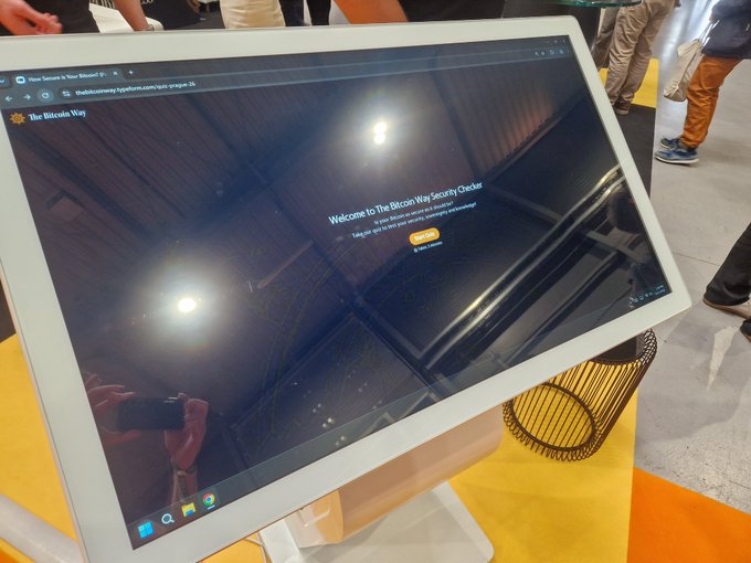 The image shows a large computer monitor with a website displayed on its screen, which appears to be about Bitcoin security. The monitor is placed on an orange table, and there are people standing around it. One person is holding a cell phone, possibly taking a picture of the monitor or looking up information related to Bitcoin security.