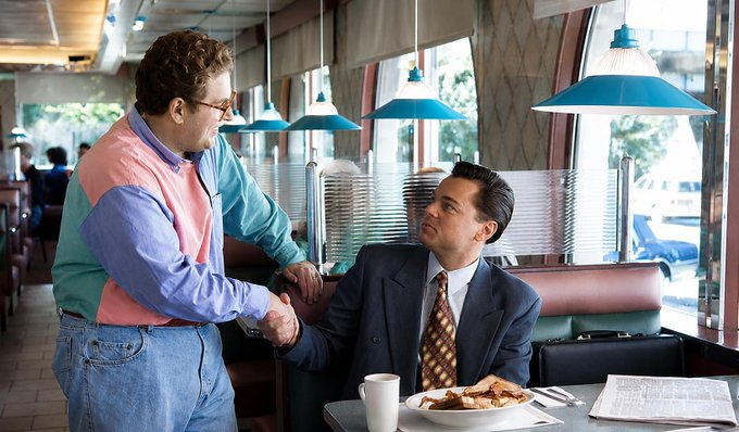 A scene from a diner showing two men in suits shaking hands, one standing in a pink and blue color-blocked shirt and the other seated at a booth with a breakfast plate.