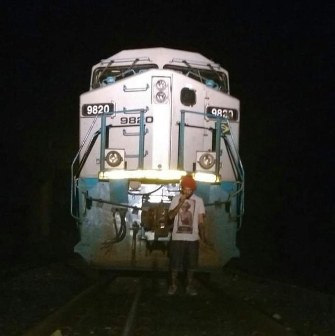 A young boy wearing a white t-shirt and red cap stands on the tracks in front of a large blue and white locomotive numbered 9820 at night.
