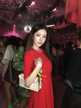 A woman in a red dress and pearl necklace holding a beige handbag at what appears to be an indoor event or party with pink and purple lighting.