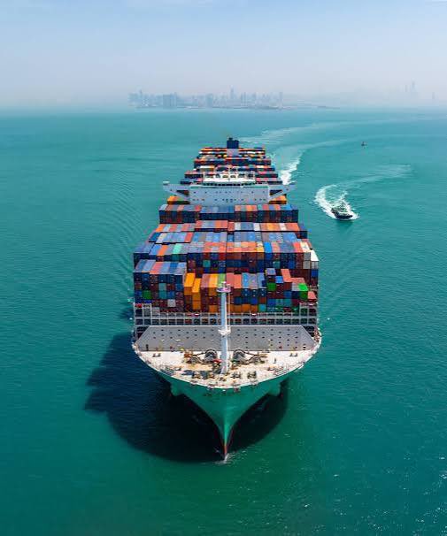 An aerial view of a massive cargo ship loaded with colorful shipping containers sailing through turquoise waters, with a small boat nearby and a city skyline in the hazy distance.