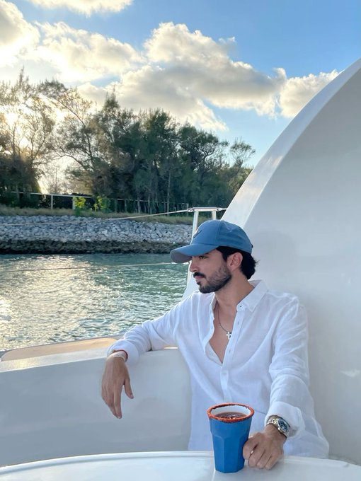 A man wearing a blue cap and white shirt sits on a white boat with a blue cup beside him, with trees and water visible in the background.