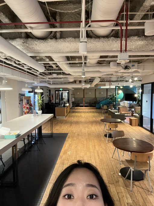 A selfie taken in a modern coworking space with exposed ceiling pipes and ductwork, featuring long communal tables, casual seating, and a blue slide visible in the background.