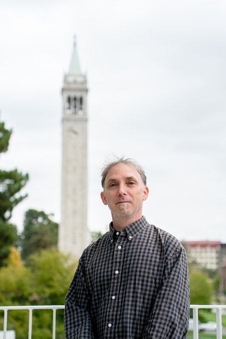 A portrait of a middle-aged man in a plaid shirt standing in front of the UC Berkeley Campanile tower on an overcast day.