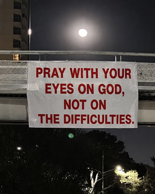 A nighttime photo of a white banner with red text reading 'PRAY WITH YOUR EYES ON GOD, NOT ON THE DIFFICULTIES.' hanging on a pedestrian bridge with the moon visible in the sky above.
