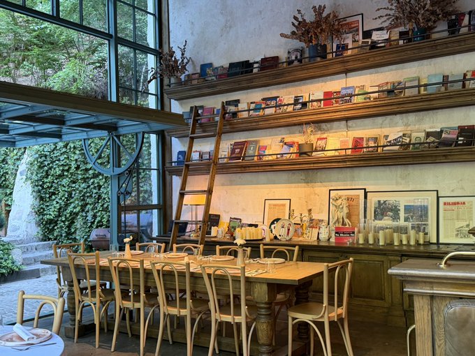A rustic dining room features a long wooden table surrounded by bentwood chairs, with shelves displaying books and magazines above a counter holding framed posters and candles, all set against a wall of large windows overlooking greenery.