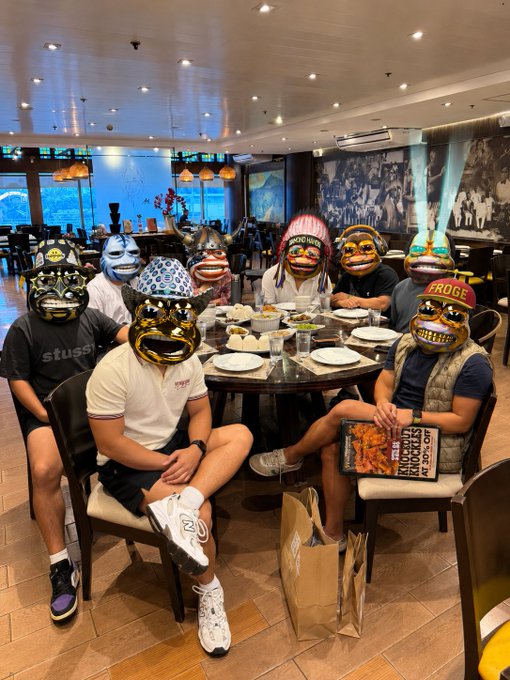 A group of people wearing colorful decorative masks sit around a dining table in a restaurant, with one person holding a 'Red Hot Chili Peppers Greatest Hits' record.