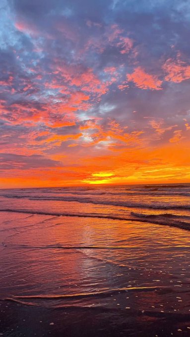 A vibrant sunset scene over the ocean, featuring orange and pink clouds reflecting on the wet sand and gentle waves.