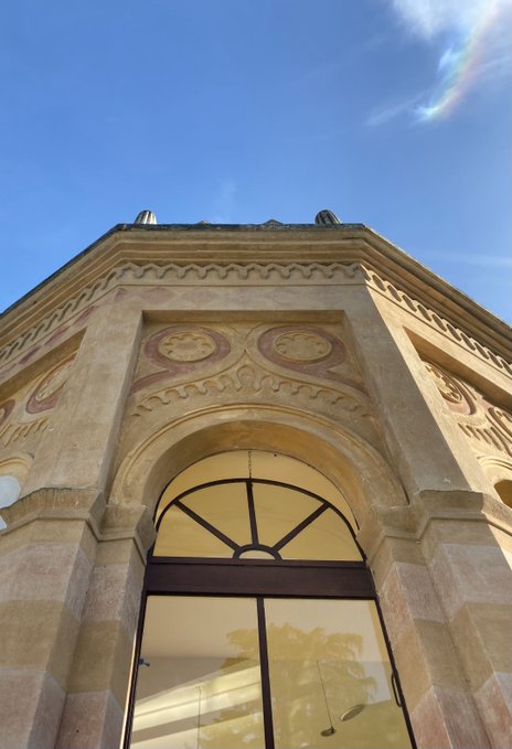 A low-angle photograph of an ornate stone building facade featuring an arched window, decorative circular motifs, and intricate carvings against a clear blue sky.