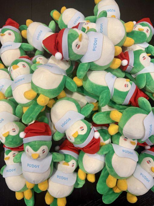 A pile of green and white penguin plush toys wearing Santa hats and scarves labeled 'PODGY'.
