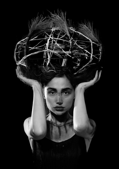 A black and white artistic portrait of a woman with a thorny, branch-like crown on her head, holding it with both hands.