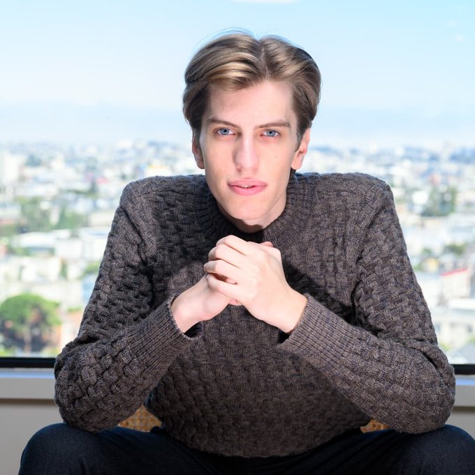 A young man with styled brown hair, wearing a thick gray knit sweater, sits with his hands clasped together in front of him against a backdrop of a sunlit city skyline visible through a window.