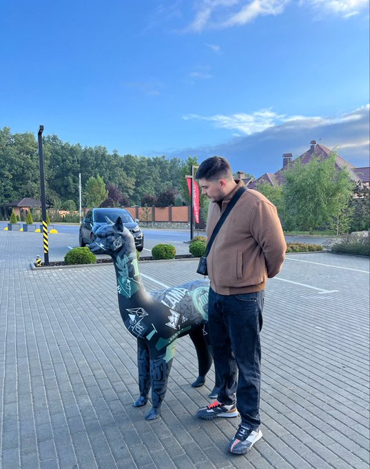 A man in a brown jacket stands on a paved area next to a black horse statue covered in graffiti-style markings.