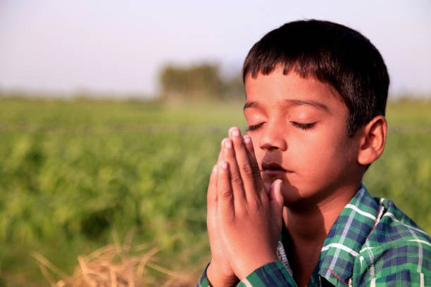 A young boy in a green plaid shirt stands with his eyes closed and hands clasped together in prayer against a backdrop of a green grassy field.