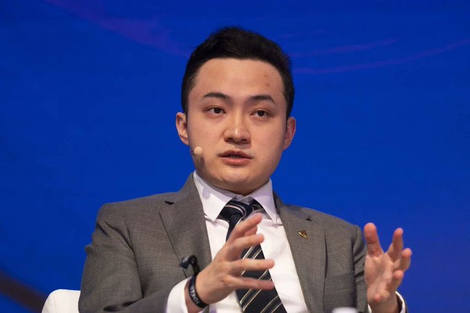 Justin Sun (TRON founder) speaking on a panel, wearing a grey suit and striped tie, gesturing with both hands against a blue background.
