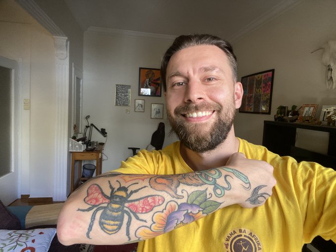 A smiling bearded man wearing a yellow shirt with 'ARMA REPUBLIC' logo, showcasing a colorful tattoo sleeve featuring a bee and floral designs on his arm, in a home interior setting.