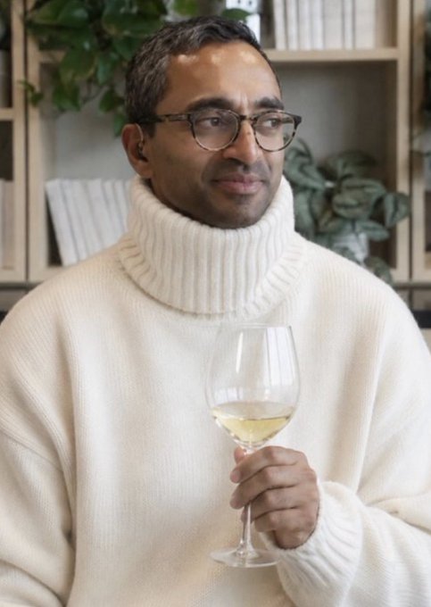 A man in a cream turtleneck sweater and round glasses holds a glass of white wine with a relaxed smile, surrounded by bookshelves and plants. The image conveys the leisurely, sophisticated lifestyle of someone who has shifted from the fast-paced grind of trading markets to appreciating life's quieter, more meaningful moments.