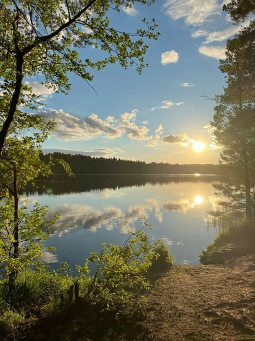 A scenic photograph captures a serene lake at sunset, with the sun reflecting on the calm water, surrounded by trees and a rocky shoreline under a partly cloudy blue sky.