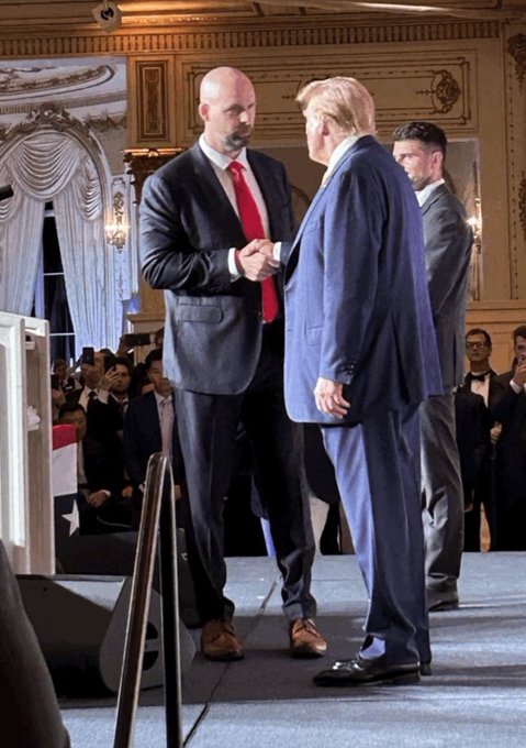 Two men in suits, one notably tall wearing a red tie, stand on a stage facing each other in what appears to be an ornate indoor venue with an audience in the background.