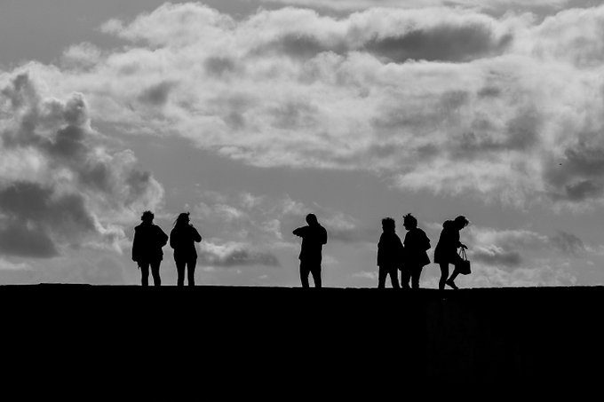 A black and white silhouette photograph of a group of people standing on a ridge against a dramatic cloudy sky.