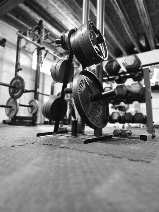 A low-angle black and white photo of a weightlifting setup in a garage gym, featuring a barbell loaded with Intek 45 lb plates and dumbbells stored on a rack in the background—showcasing the gritty, no-frills training space that earns "mad respect" for its dedication to the grind.