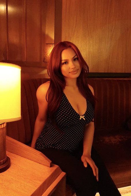 A red-haired woman sits on a brown couch in a dimly lit room, wearing a black top with white polka dots and a small bow, with a warm lamp glowing on a side table beside her.