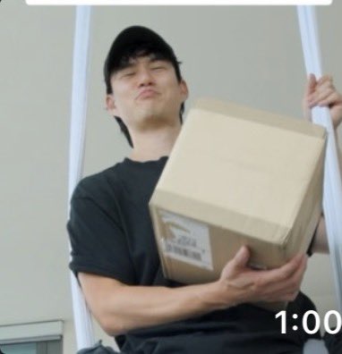 A strained young man holds a large cardboard package with a grimacing expression, paired with text displaying "1:00," visually capturing the physical toll implied by "roundtripped 6 figures today."