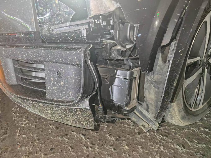 A damaged black car shows significant front-end collision damage with the bumper and headlight area broken off, covered in mud and debris.