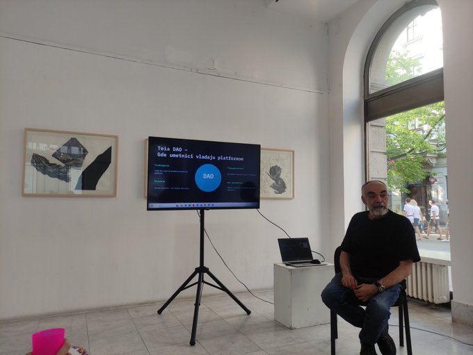 A man with a gray beard and black shirt sits on a stool in a gallery-like space next to a TV screen on a tripod displaying a presentation titled 'Trečia DAO' and 'Gde umetnost vladaju platformom' with a large blue 'DAO' logo.