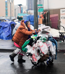 A person in an orange puffer jacket pushes a cart overflowing with white plastic bags labeled 'Verde' through a rainy urban street lined with blue tarps and stacked boxes.